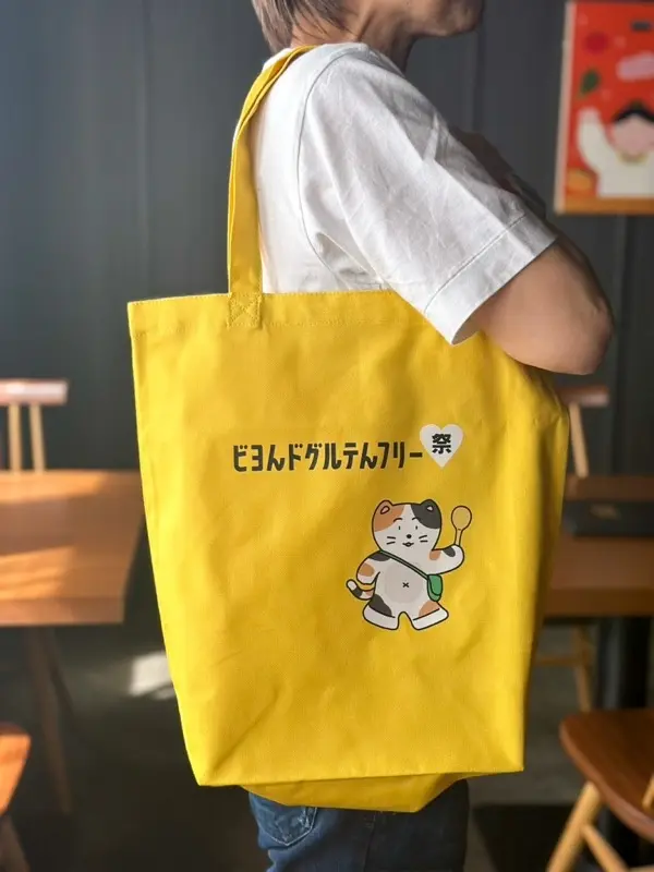黄色の応援トートバッグを肩に掛けた様子。猫のキャラクターとロゴ入り